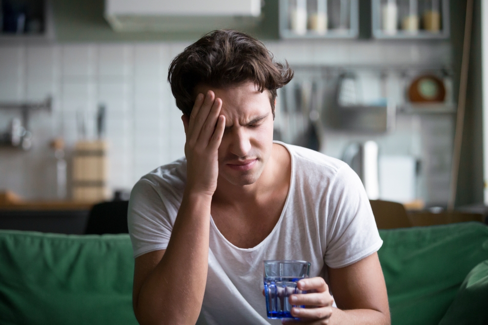 man holding his head with his hands due to his headache while holding a glass of water